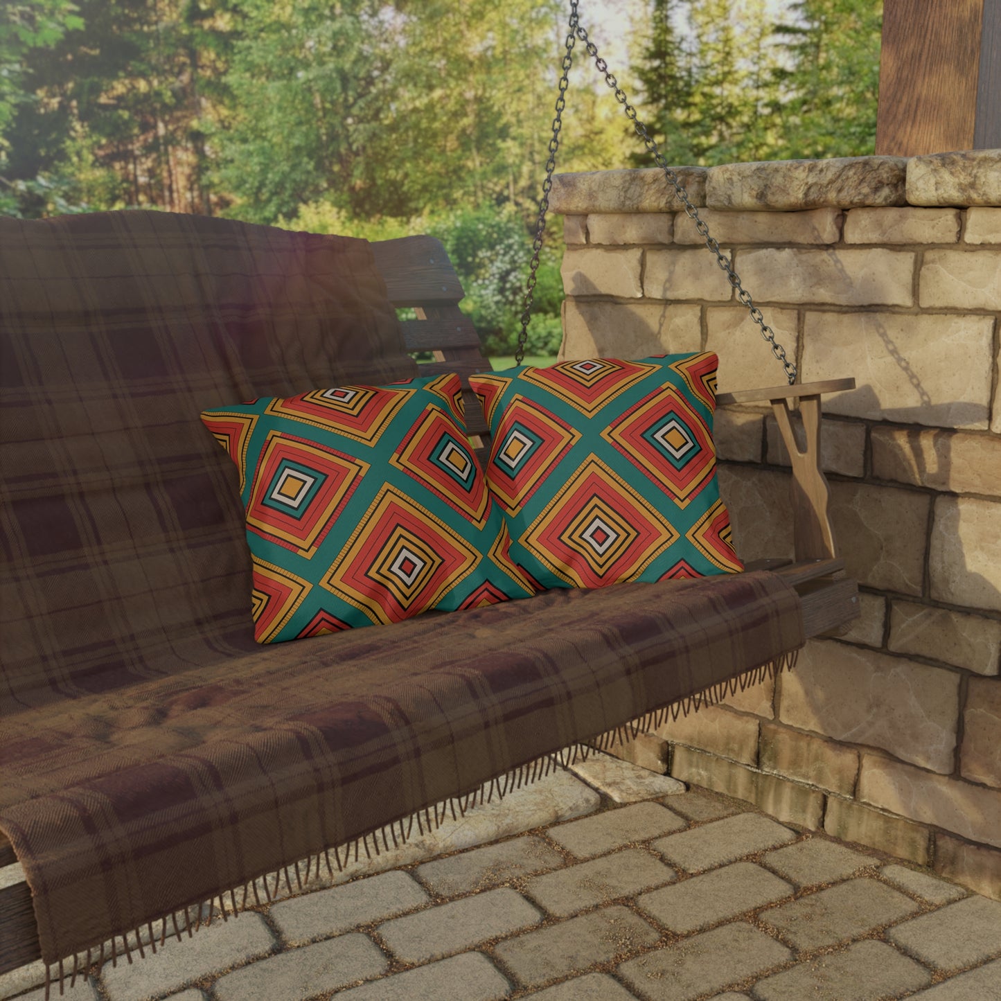 A wooden porch swing with a plaid blanket and two colorful outdoor pillows hangs from chains beside a stone wall. The swing sits on a stone patio, surrounded by greenery and trees, creating an inviting spot for sunny days.