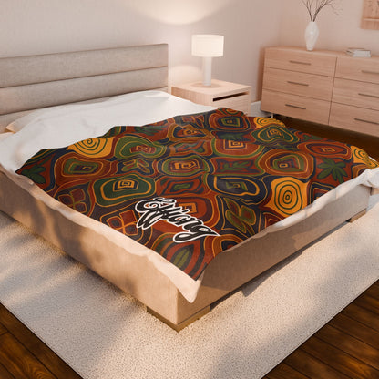 A neatly made bed with a colorful, African-inspired blanket in warm shades of red, orange, yellow, and green, featuring a white script logo in one corner. The room has a beige headboard, wooden furniture, white lamp, and light décor.