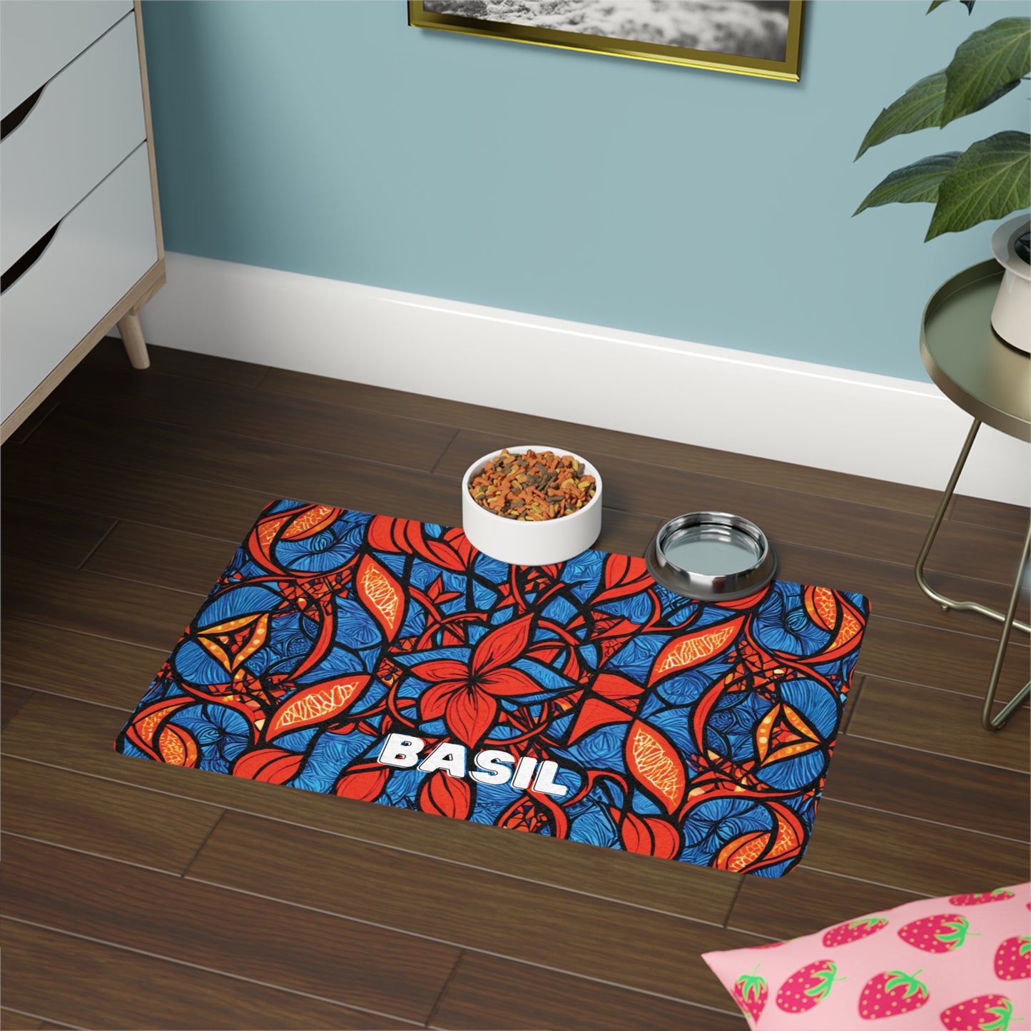 A vibrant African-inspired pet mat with red, orange, and blue leaf patterns and the name “BASIL” in bold white letters sits on a wood floor. A food bowl with kibble and a metal water bowl rest on the mat. Nearby are a plant, dresser, and strawberry-patterned cloth.
