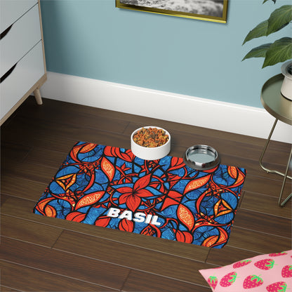A vibrant African-inspired pet mat with red, orange, and blue leaf patterns and the name “BASIL” in bold white letters sits on a wood floor. A food bowl with kibble and a metal water bowl rest on the mat. Nearby are a plant, dresser, and strawberry-patterned cloth.