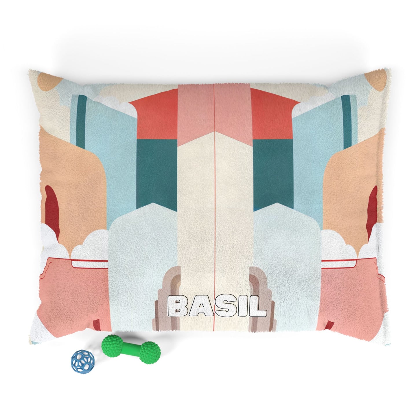 A soft rectangular pet bed features an African-inspired abstract geometric pattern in muted red, blue, beige, and brown tones with “BASIL” written boldly at the center. In front are a green dumbbell-shaped toy and a blue ball toy.