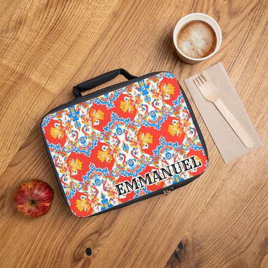A colorful, African-inspired personalized lunch bag with a red, yellow, blue, and white floral pattern and the name EMMANUEL sits on a wooden table next to a red apple, coffee, a brown napkin, and wooden utensils.