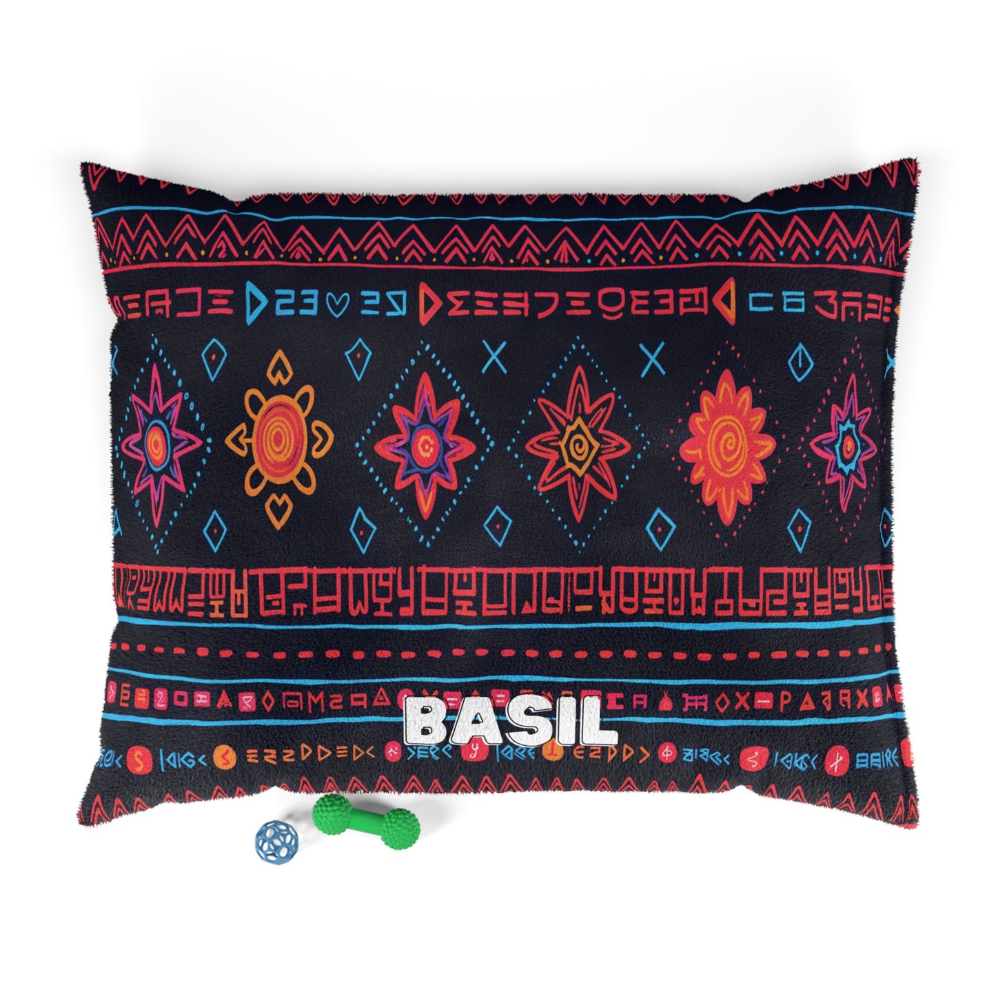 A rectangular pillow featuring colorful red, orange, and blue geometric and star patterns on a black background offers luxurious comfort. “BASIL” is printed in white block letters. Nearby are a green textured bone toy and a blue ball.