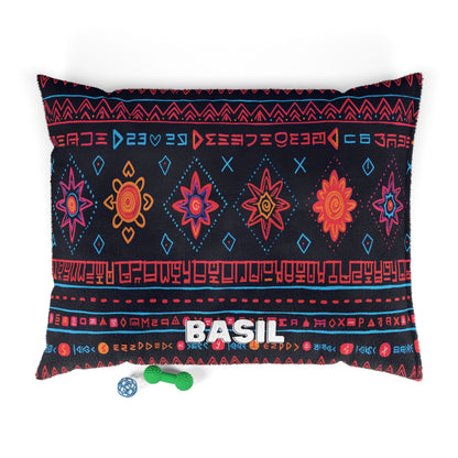 A rectangular pillow featuring colorful red, orange, and blue geometric and star patterns on a black background offers luxurious comfort. “BASIL” is printed in white block letters. Nearby are a green textured bone toy and a blue ball.