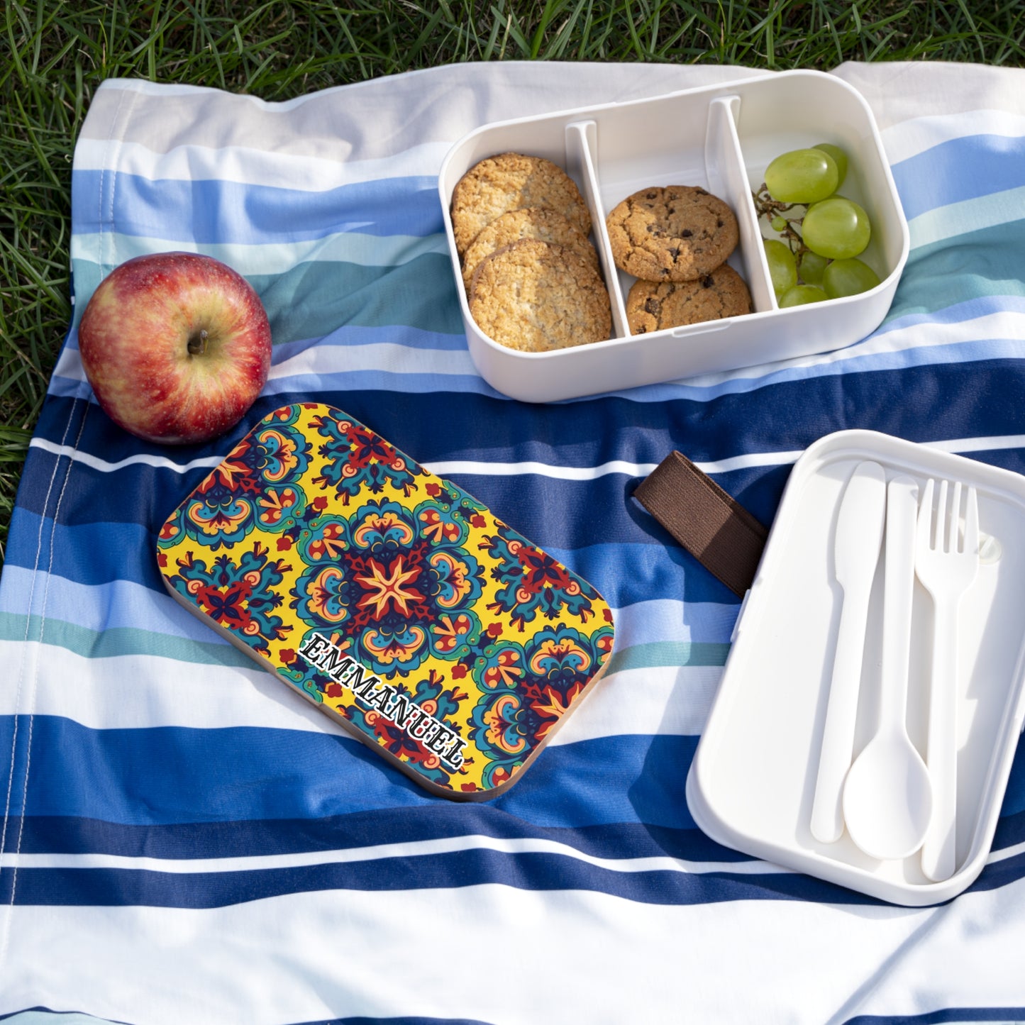 A colorful, BPA-free personalized lunch box with a floral pattern and “EMMANUEL” label sits on a blue striped picnic blanket. Nearby are containers of cookies, green grapes, white cutlery, and an apple resting on the grass.