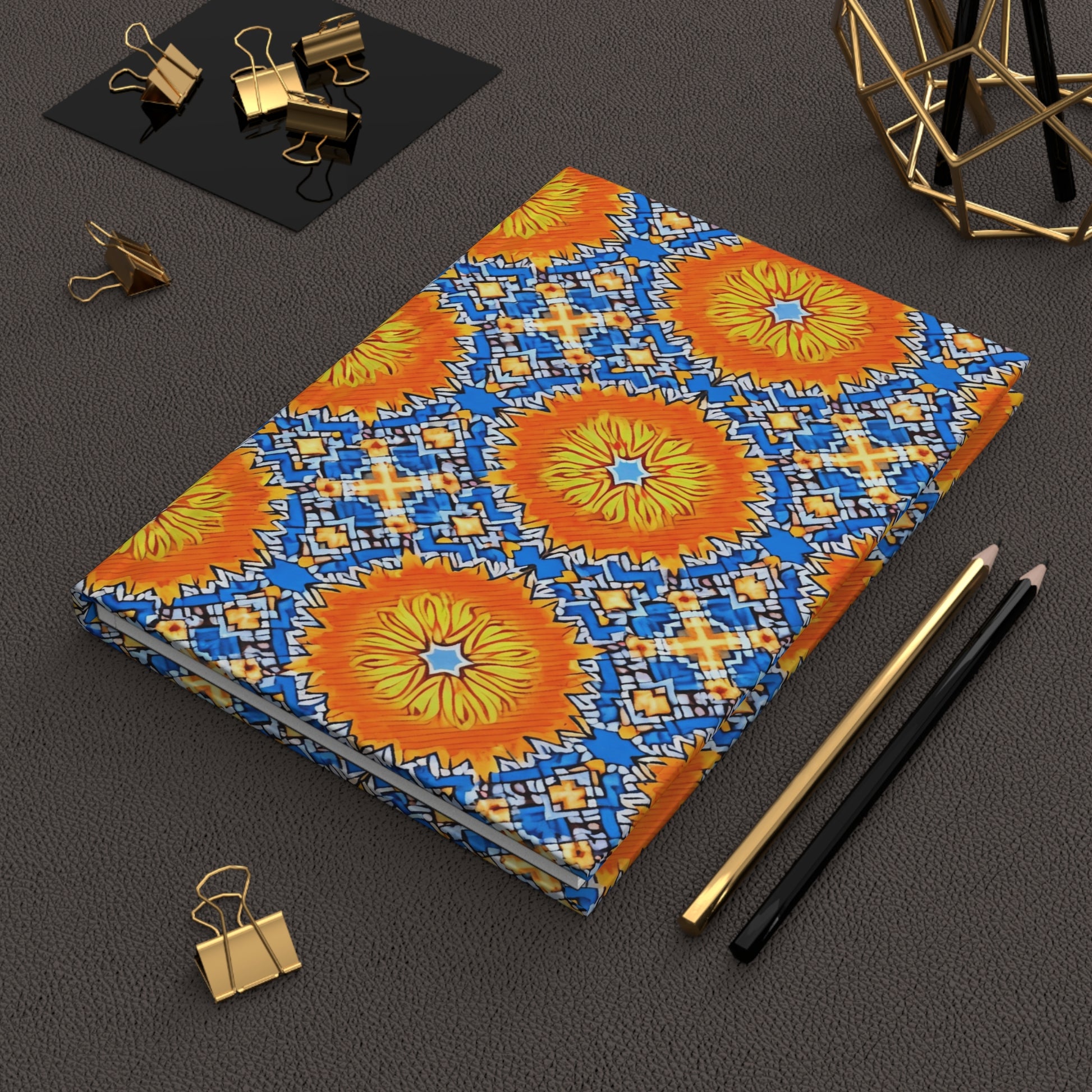 A vibrant African-inspired hardcover journal with a geometric pattern in blue, orange, and yellow lies on a textured brown surface near two pencils, gold binder clips, a black pad, and a decorative gold wire sculpture.