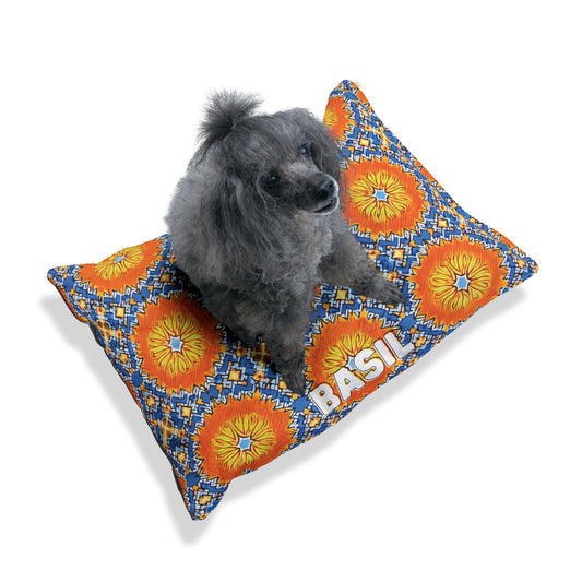 A small gray poodle with fluffy fur sits on a vibrant African inspired pet bed. Featuring orange and yellow sunbursts, blue accents, and BASIL in bold white letters, this personalized pet bed stands out against the crisp white background.