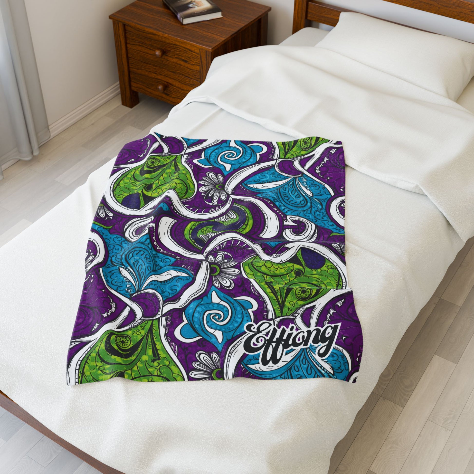 A single bed with white bedding and a wooden nightstand beside it. A personalized velveteen blanket featuring green, blue, and purple abstract paisley patterns and the word Effing is spread across the lower half of the bed.