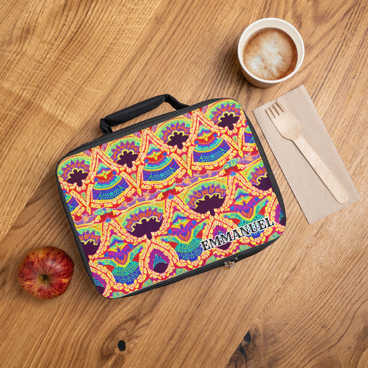 A colorful, African-inspired lunch tote with a vibrant geometric pattern and the text “EMMANUEL” sits on a wooden table next to a red apple, frothy coffee, and wooden cutlery on a napkin.