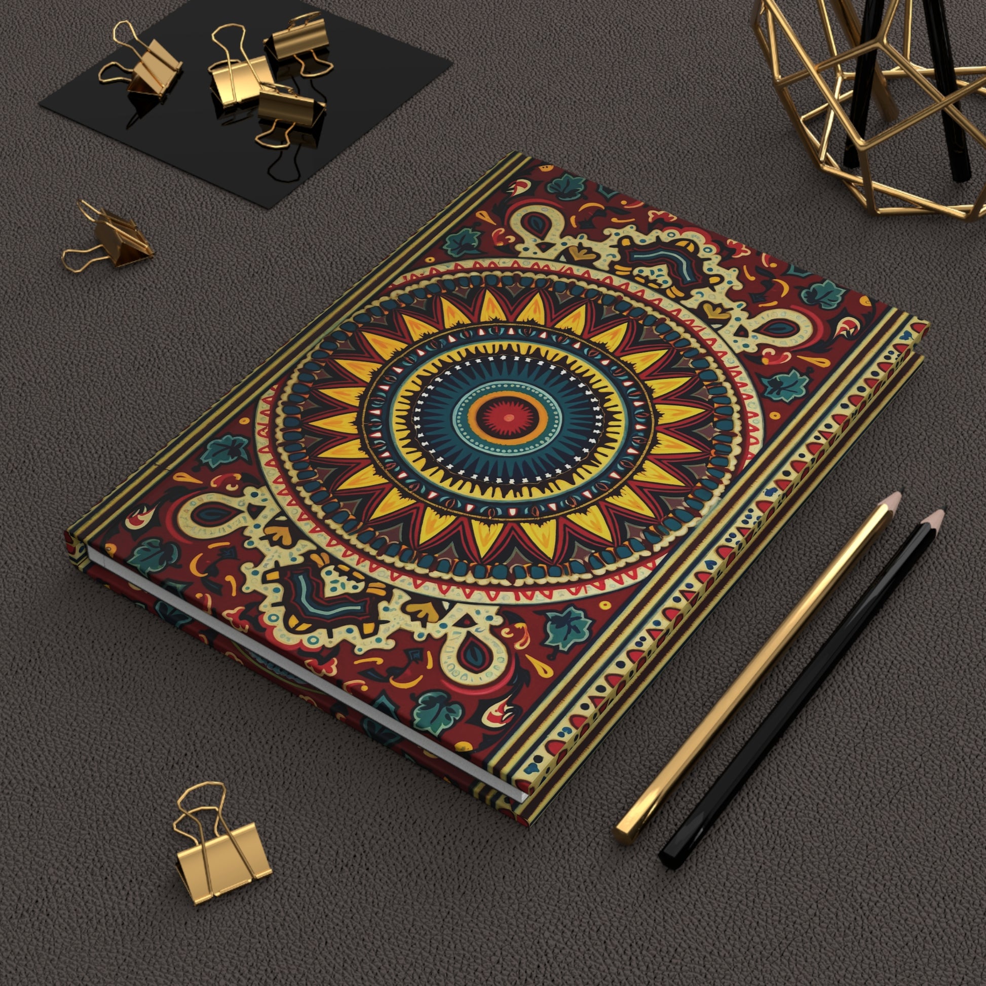 A colorful hardcover journal with an intricate mandala design lies on a textured brown surface, flanked by a gold pencil, a black pencil, binder clips, and a geometric gold desk ornament in the upper right corner.