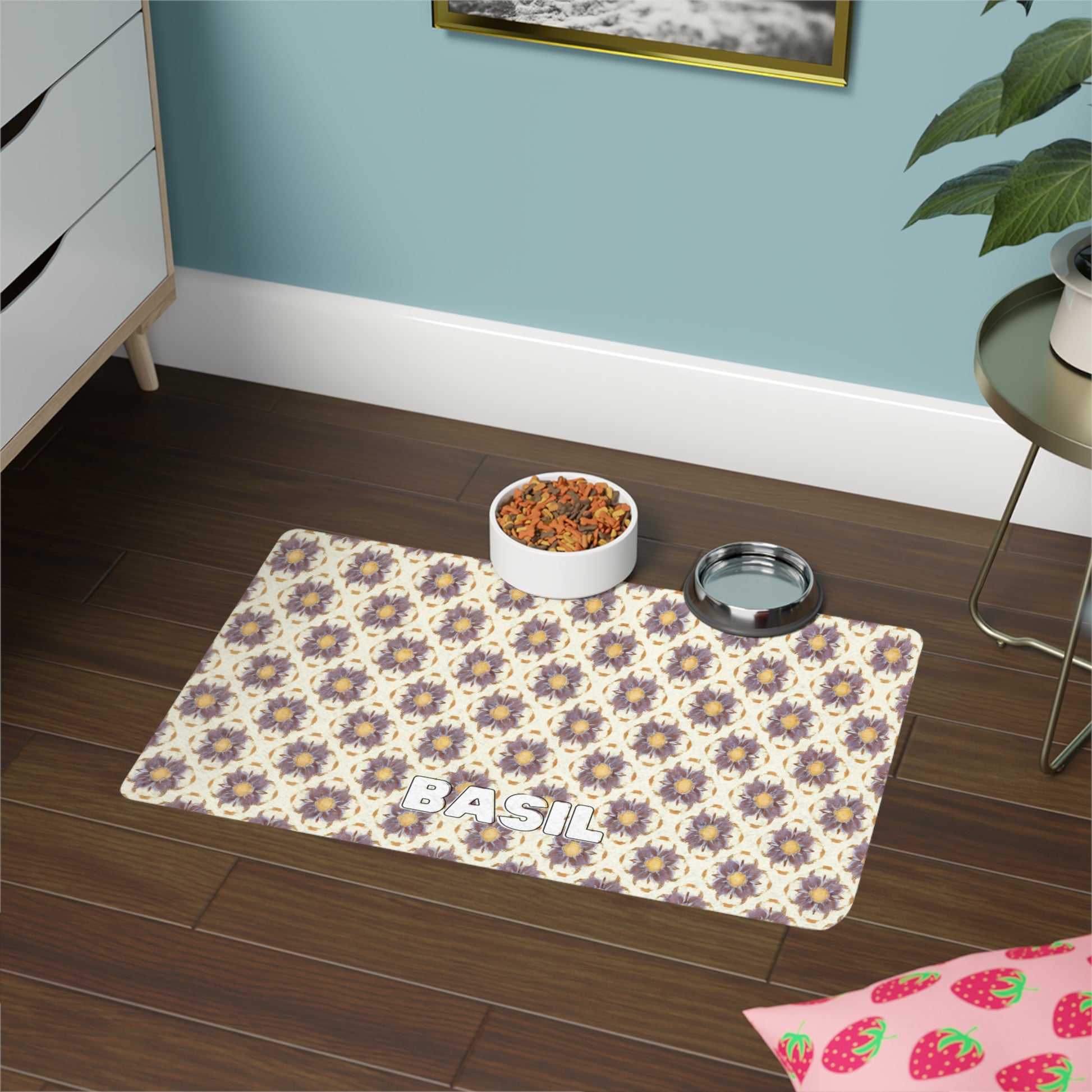 A patterned, African-inspired pet mat with “BASIL” written on it sits on a brown wooden floor, holding a bowl of kibble and a metal water dish. Nearby are drawers, a green plant, blue walls, and a pink strawberry-printed cushion in the corner.