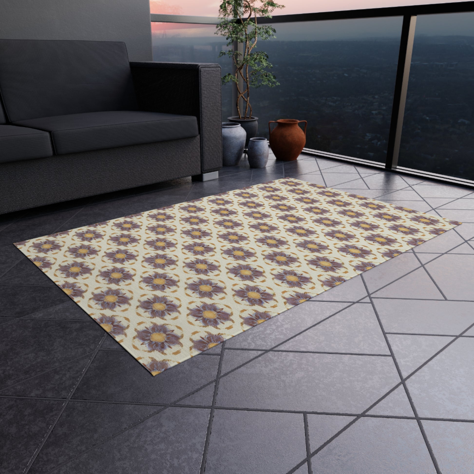 A balcony with gray tile floor features an African-inspired rectangular rug with purple and yellow floral designs. A dark gray sofa, several clay pots, and a leafy plant sit near a glass railing overlooking a cityscape at sunset.