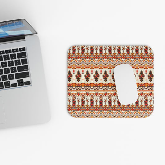 A silver laptop sits partially open on a white surface beside a vibrant desk accessory—an African-inspired mouse pad with a non-slip rubber base, its intricate orange, brown, and cream pattern topped with a white wireless mouse. The overall scene is minimalist and tidy.