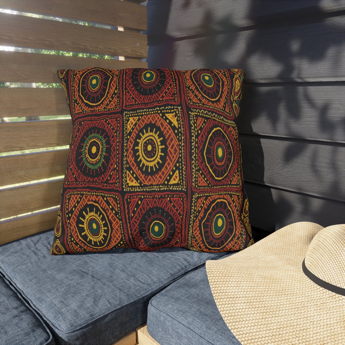 A square pillow with a bold, African-inspired geometric red, yellow, and green tribal pattern rests on a blue outdoor bench. Sunlight casts leafy shadows on a wooden fence and dark horizontal siding behind it. Part of a straw hat appears nearby.