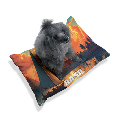 A small gray poodle with curly fur sits on a personalized pet bed—a pillow printed with an orange sunset, palm trees, a sailboat, and BASIL in bold letters. The plain white background highlights the luxurious comfort of this unique pet accessory.