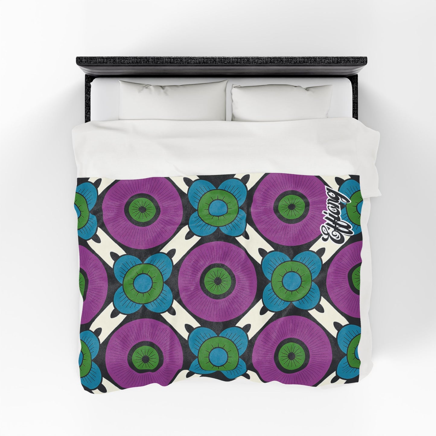 A bed with a velveteen blanket featuring a bold floral pattern of large purple and blue flowers with green centers, arranged in a diamond grid on cream. The word “Enjoy” appears in the lower right corner. Two white pillows rest atop this customizable gift.