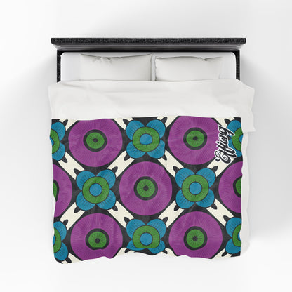 A bed with a velveteen blanket featuring a bold floral pattern of large purple and blue flowers with green centers, arranged in a diamond grid on cream. The word “Enjoy” appears in the lower right corner. Two white pillows rest atop this customizable gift.
