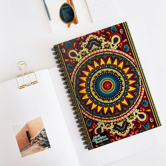 A vibrant designs journal with an intricate mandala in red, yellow, blue, and green rests on a white desk. Nearby are sunglasses, a gold paper clip, a notepad, and an open magazine showing an orange wall and stairs.
