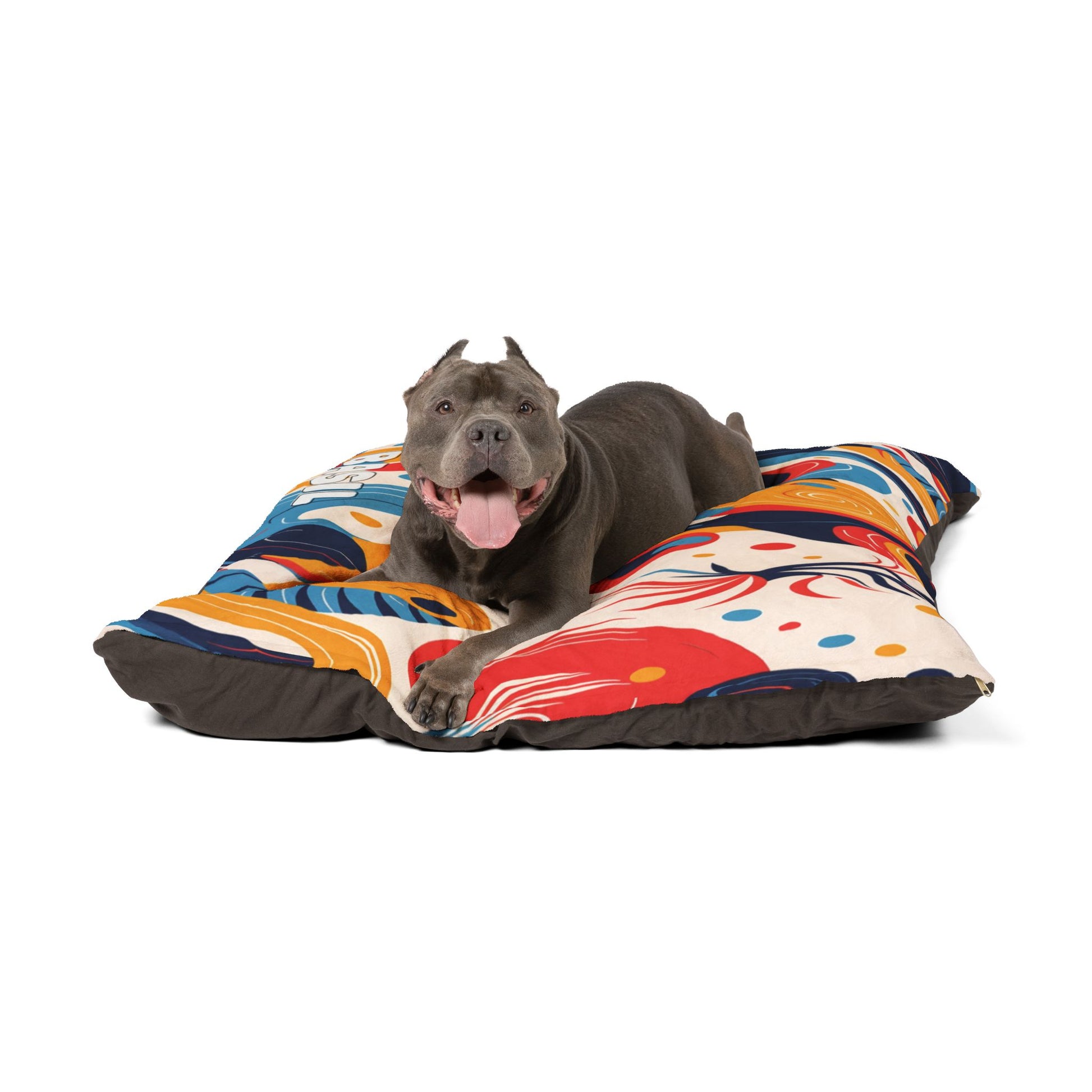 A happy gray pit bull with cropped ears lies on a colorful, African-inspired dog bed featuring abstract patterns in red, blue, yellow, and orange. The dog has its mouth open and tongue out, looking forward against a plain white background.