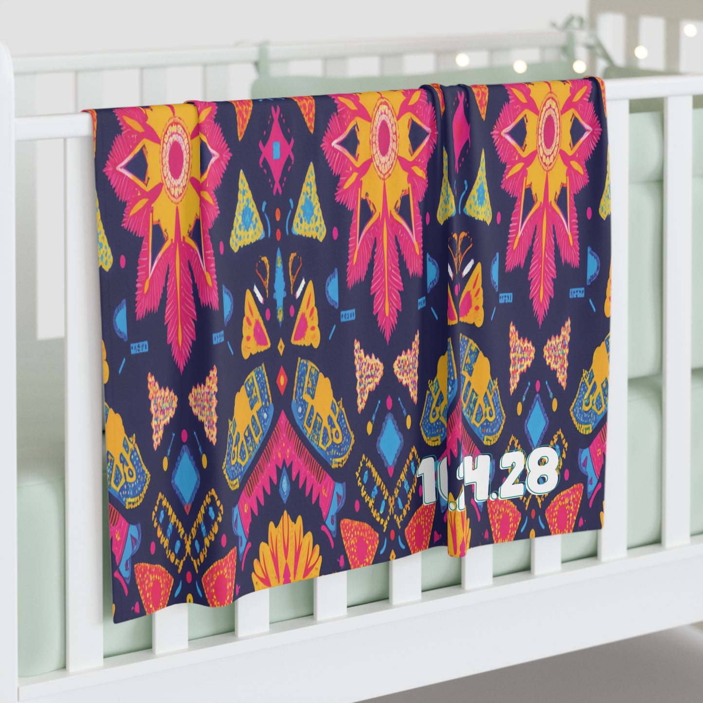 A colorful personalized baby blanket with bright orange, yellow, pink, and blue geometric floral patterns hangs over a white crib. The numbers 11.14.28 are printed in white on the lower right side. The crib bedding is light mint green.