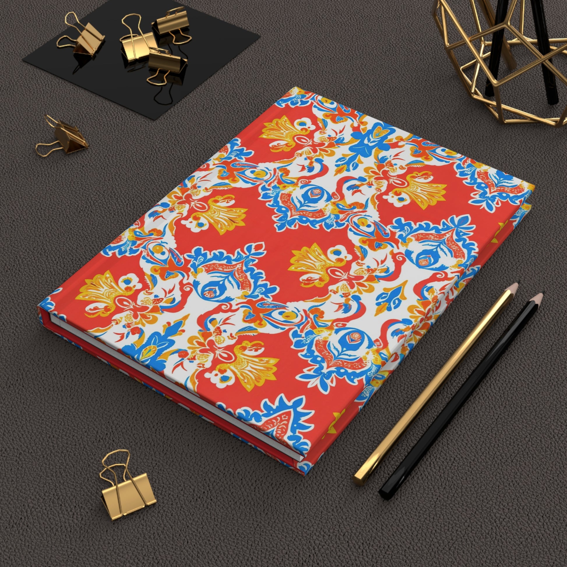 A colorful African-inspired notebook with a red, blue, yellow, and white ornate floral pattern lies on a dark textured desk. Nearby are a gold and black pencil, gold geometric decor, and gold binder clips—some on a black tray and one loose.