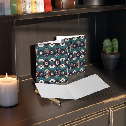 A patterned greeting card with geometric circles and a culturally inspired design stands open on a dark wooden desk, next to an open blank card, a candle, pen, bookshelf above, and a small potted cactus. Warm light creates a cozy atmosphere.