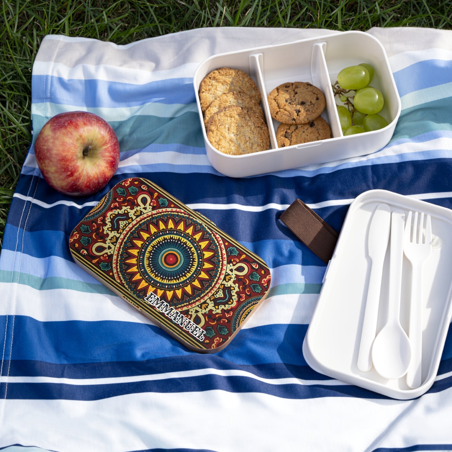 A colorful, BPA-free lunchbox with a mandala design sits on a striped picnic blanket beside a divided container holding cookies and grapes, an apple, and white cutlery in a case. The scene is outdoors on grass.