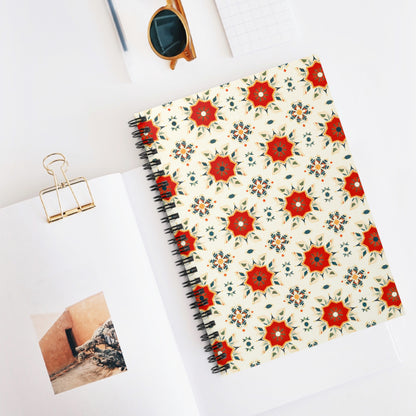 A vibrant African-inspired spiral notebook with a red and orange floral geometric pattern lies on a white desk, beside an open book with a photo of a peach wall, a gold binder clip, sunglasses, and a small grid notepad.