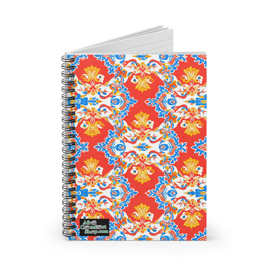 A spiral-bound African-inspired spiral notebook with a vibrant red cover, intricate yellow, blue, and white floral patterns, and an “Aim Confident Shop.com” logo at the bottom left. The notebook is partially open to reveal premium writing paper.