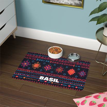 A colorful, African-inspired geometric-patterned pet mat labeled BASIL holds two bowls—one with dry food, one metal and empty. It sits on a dark wooden floor near a white cabinet, green plant, strawberry-print pillow, and framed photo.