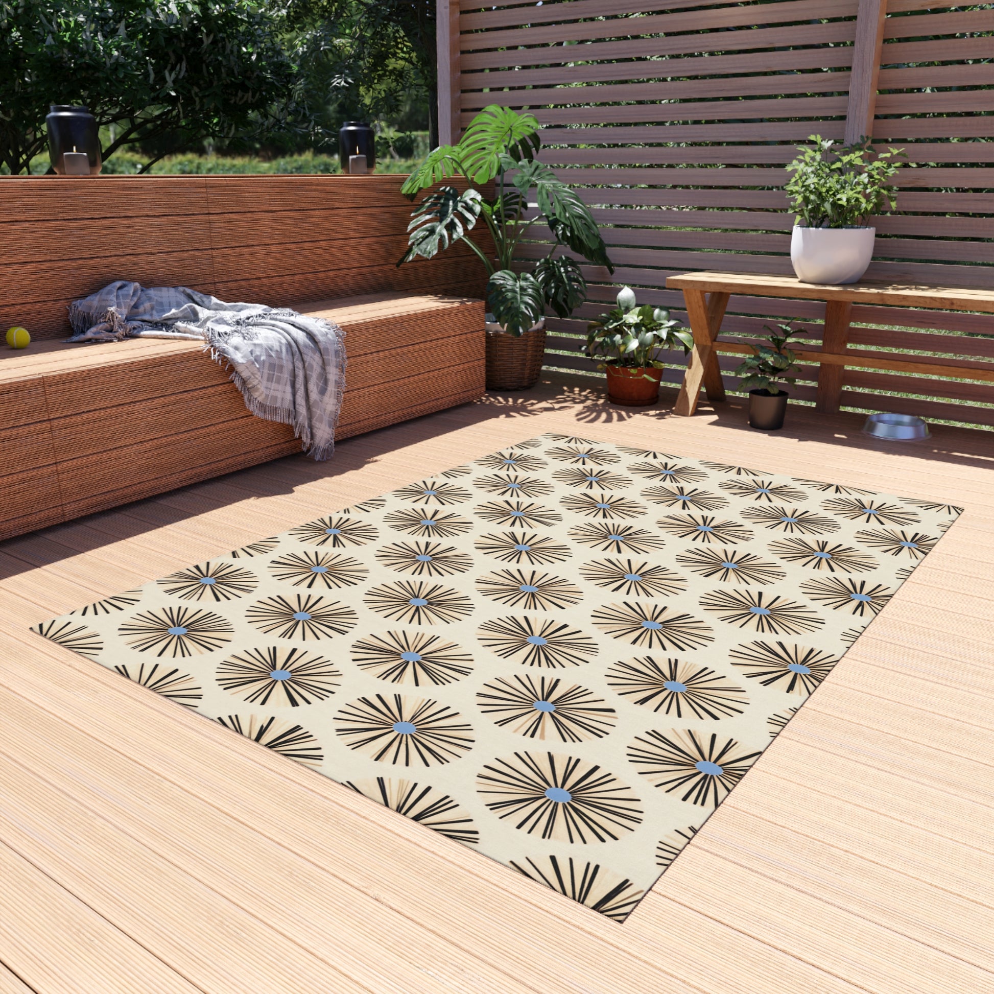 A patio with a tan weather-resistant polyester chenille rug featuring black starburst patterns inspired by authentic African art designs, a wicker sofa with a gray throw, green plants, and a wooden table. Sunlight filters through the slatted privacy screen and trees.