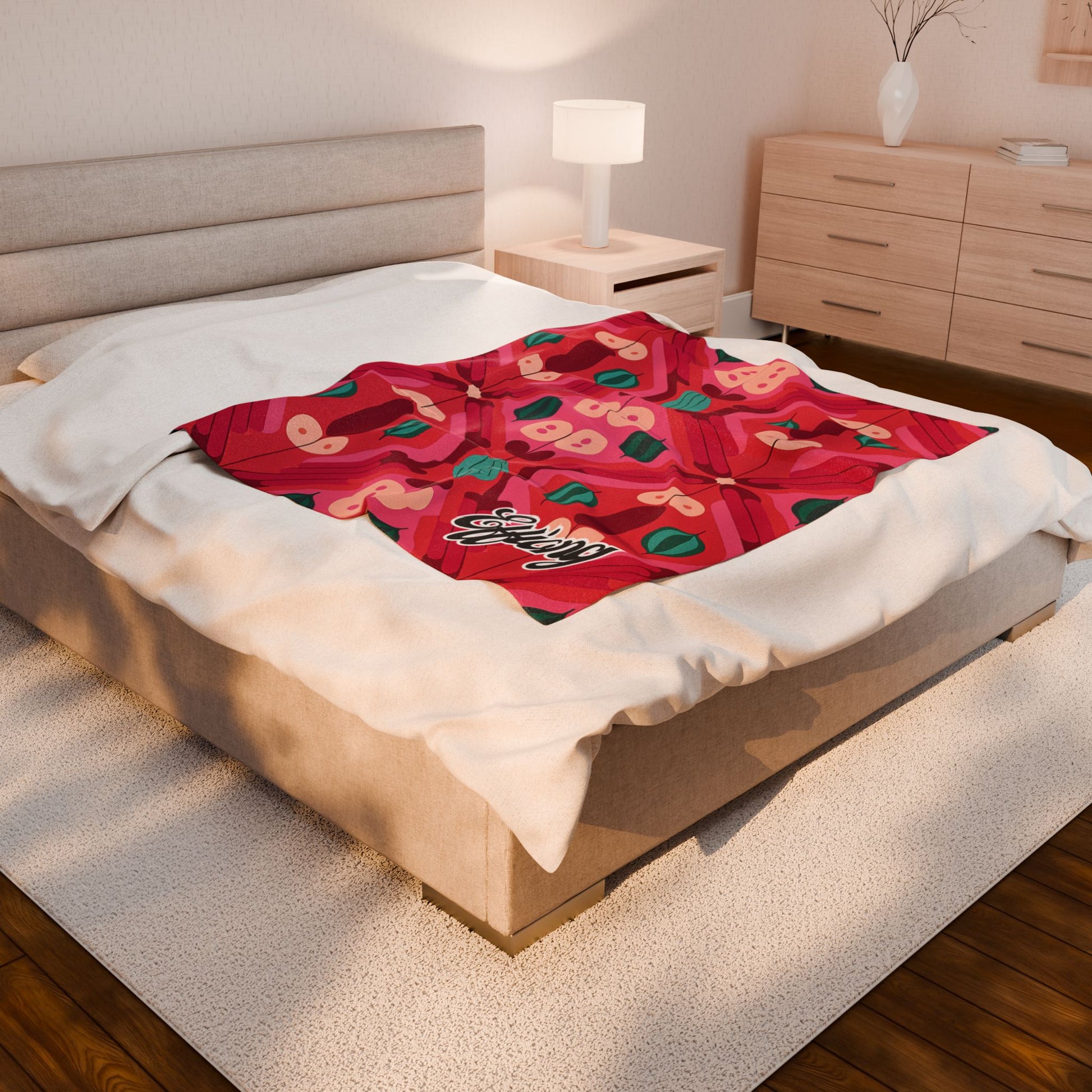 A cozy beige bed with a soft white comforter is topped with a vibrant red personalized velveteen blanket featuring playful apple and leaf patterns. The blanket has “Enjoy” written in white script. The bedroom has wooden furniture, warm lighting, and minimalist decor.