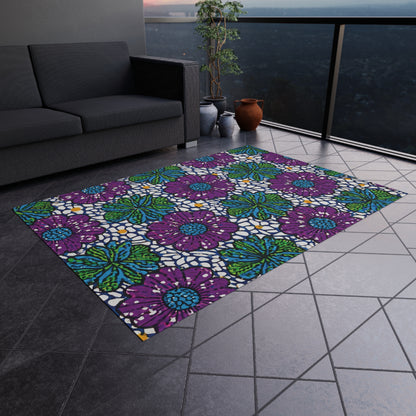 A colorful polyester chenille rug with bold purple flowers, blue centers, and green leaves on white brightens a tiled balcony floor beside a dark gray sofa, potted plants, and brown vases, with a cityscape visible beyond the glass railing.