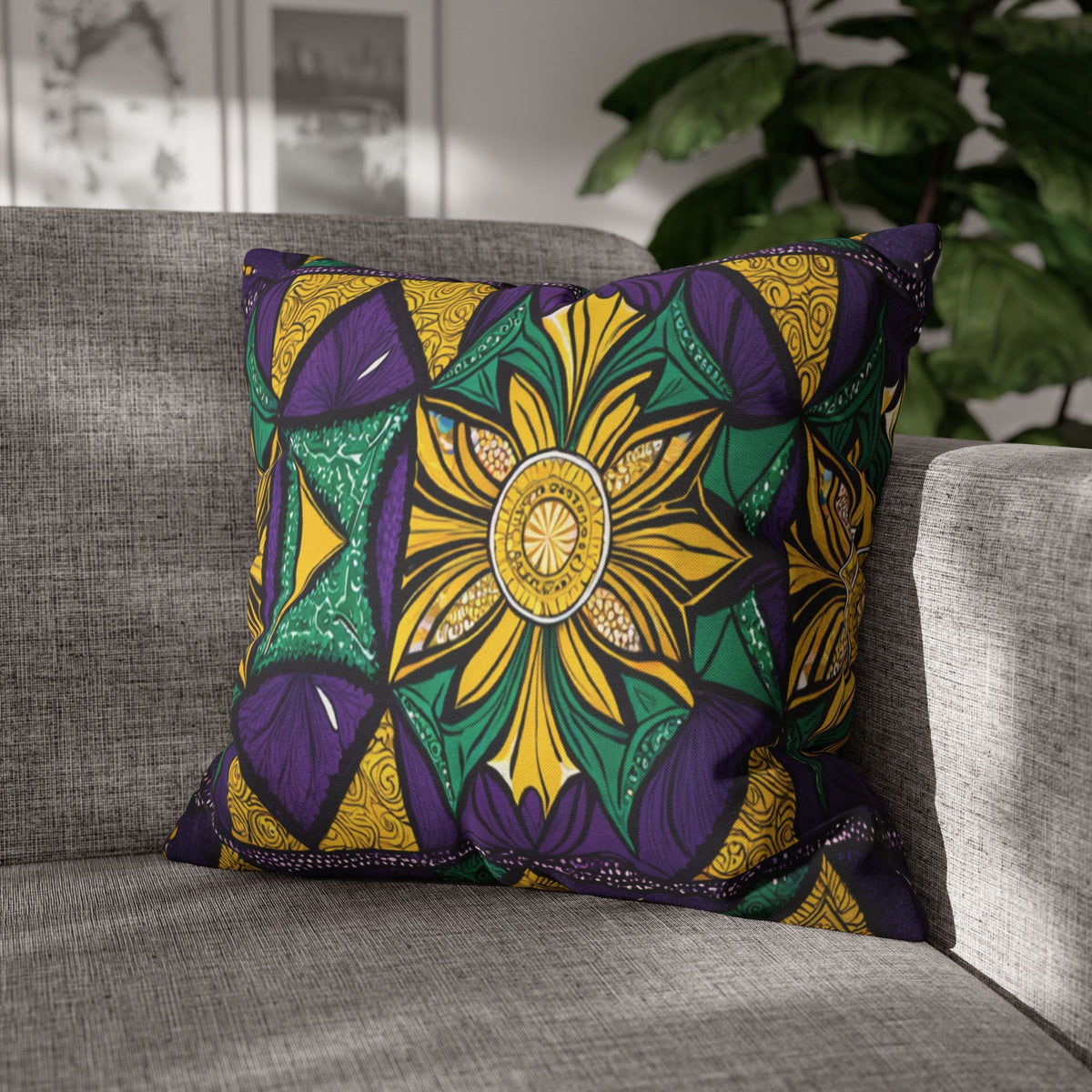 A square pillow with bold African patterns featuring a large yellow flower at the center, surrounded by green, purple, and yellow geometric shapes, sits on a light gray sofa. A leafy plant and framed art are visible in the blurred background.