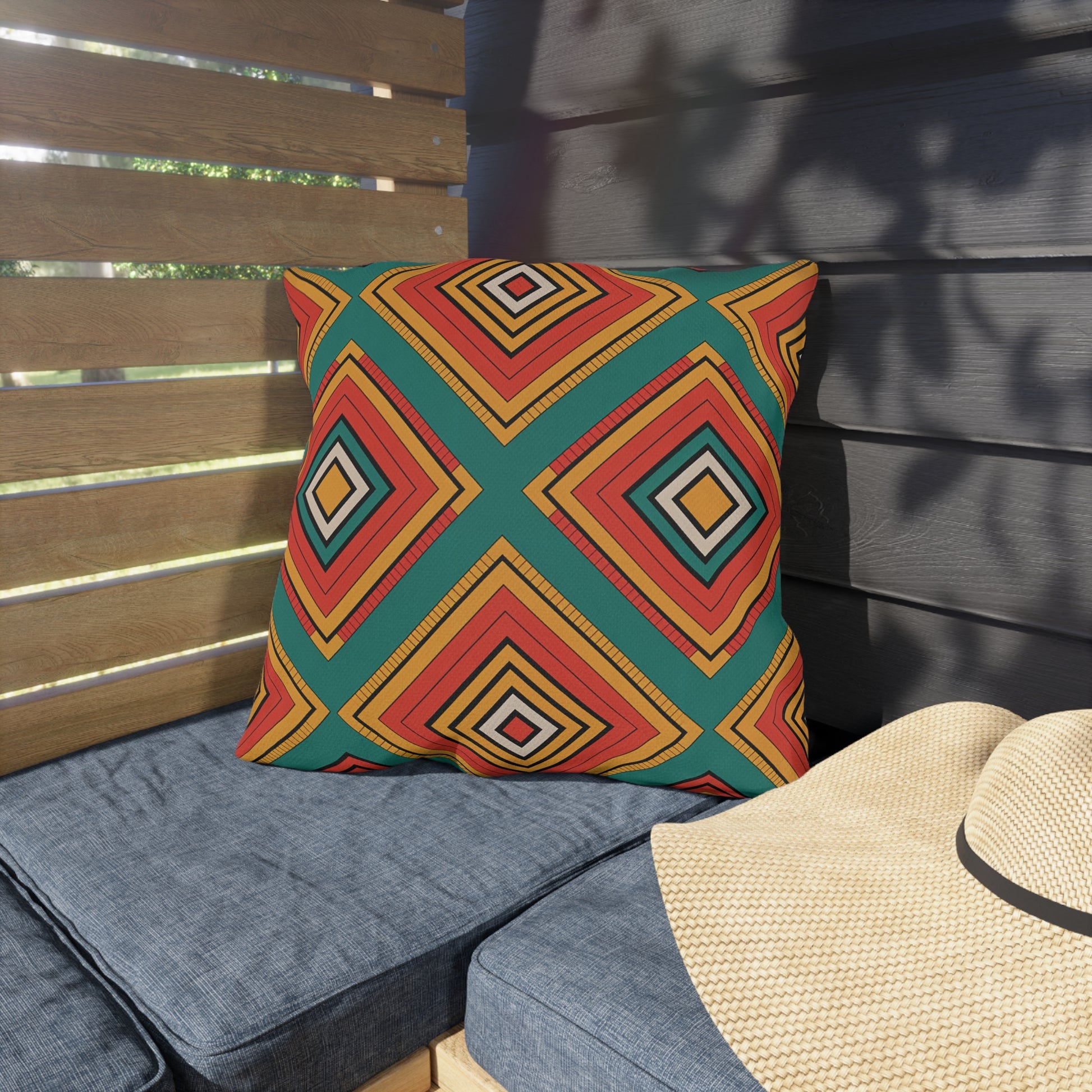 A colorful outdoor pillow with a bold geometric diamond pattern in red, yellow, orange, and green rests on a gray cushioned bench. Made from weatherproof materials, it sits beside a woven straw hat as sunlight creates patterned shadows in the background.