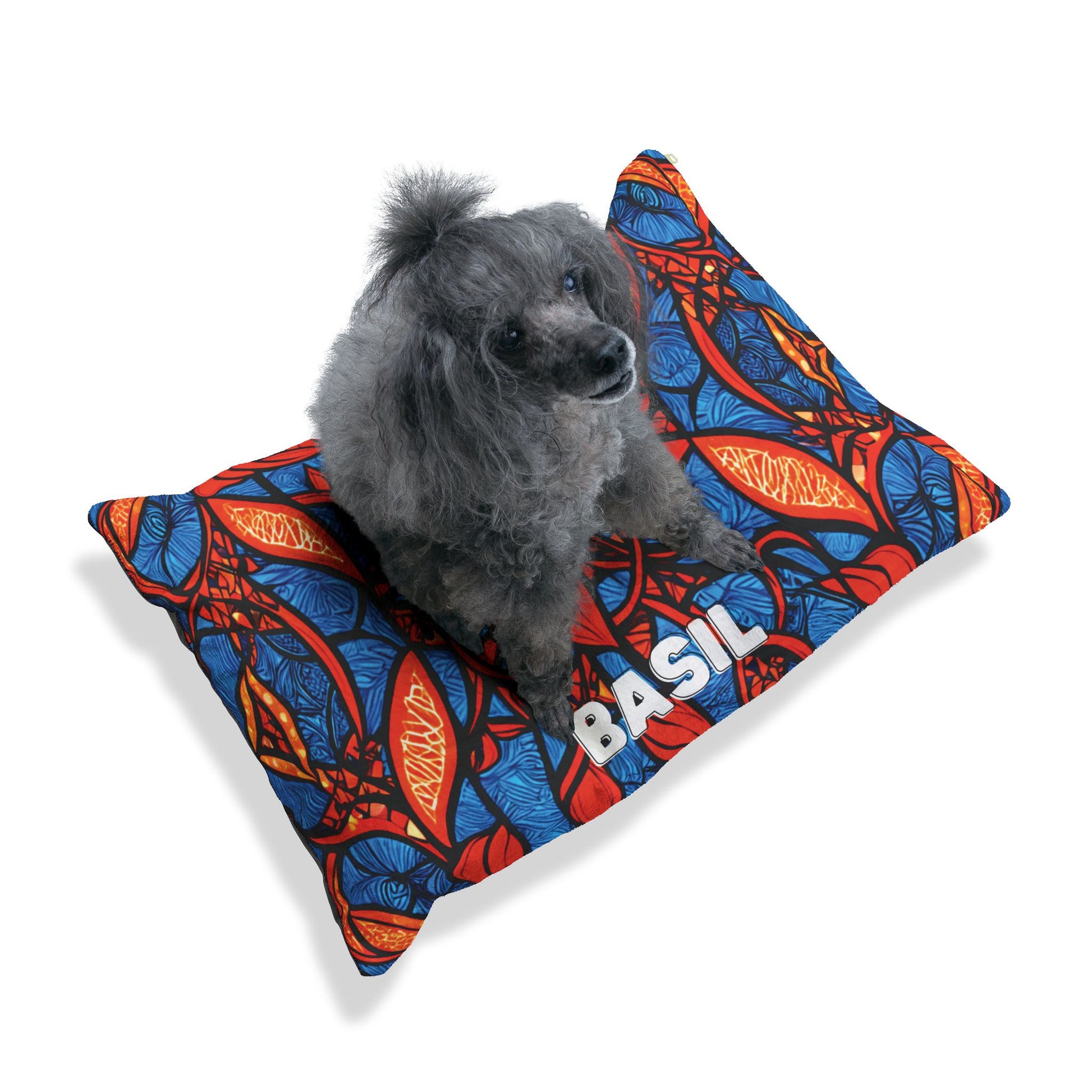 A small gray poodle sits on a cozy personalized pet bed—a colorful pillow with a bold red, blue, and orange abstract leaf pattern. The name BASIL is printed in large white letters. The background is plain white with a soft shadow under the pillow.