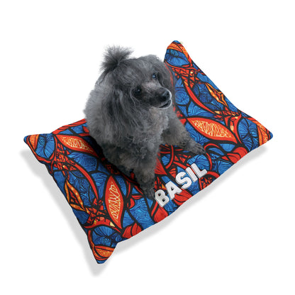 A small gray poodle sits on a cozy personalized pet bed—a colorful pillow with a bold red, blue, and orange abstract leaf pattern. The name BASIL is printed in large white letters. The background is plain white with a soft shadow under the pillow.