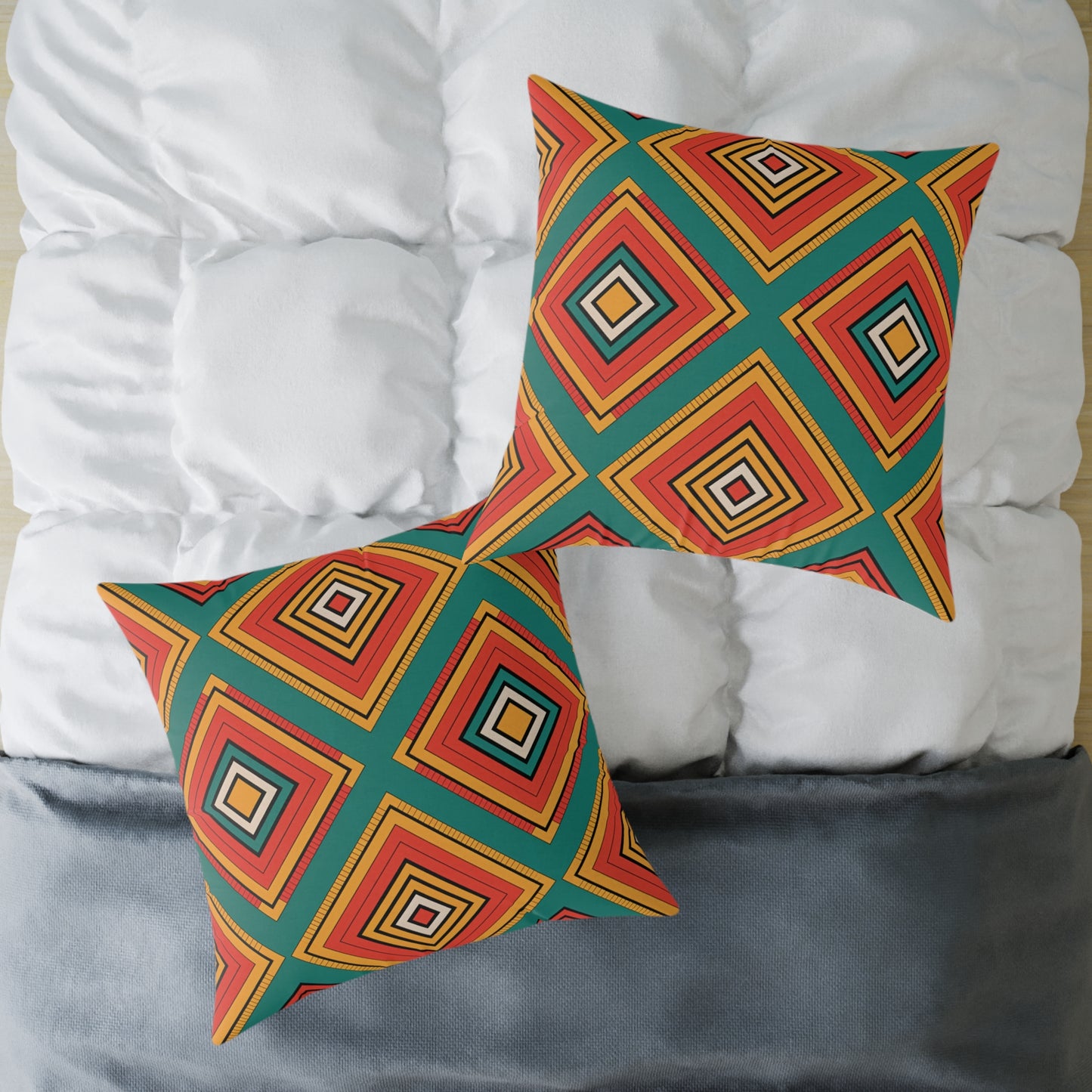 Two square African-inspired pillows with vibrant geometric diamond patterns in orange, yellow, red, and teal rest on a neatly made bed. The white quilted comforter and gray blanket highlight these colorful home decor accents arranged at an angle.