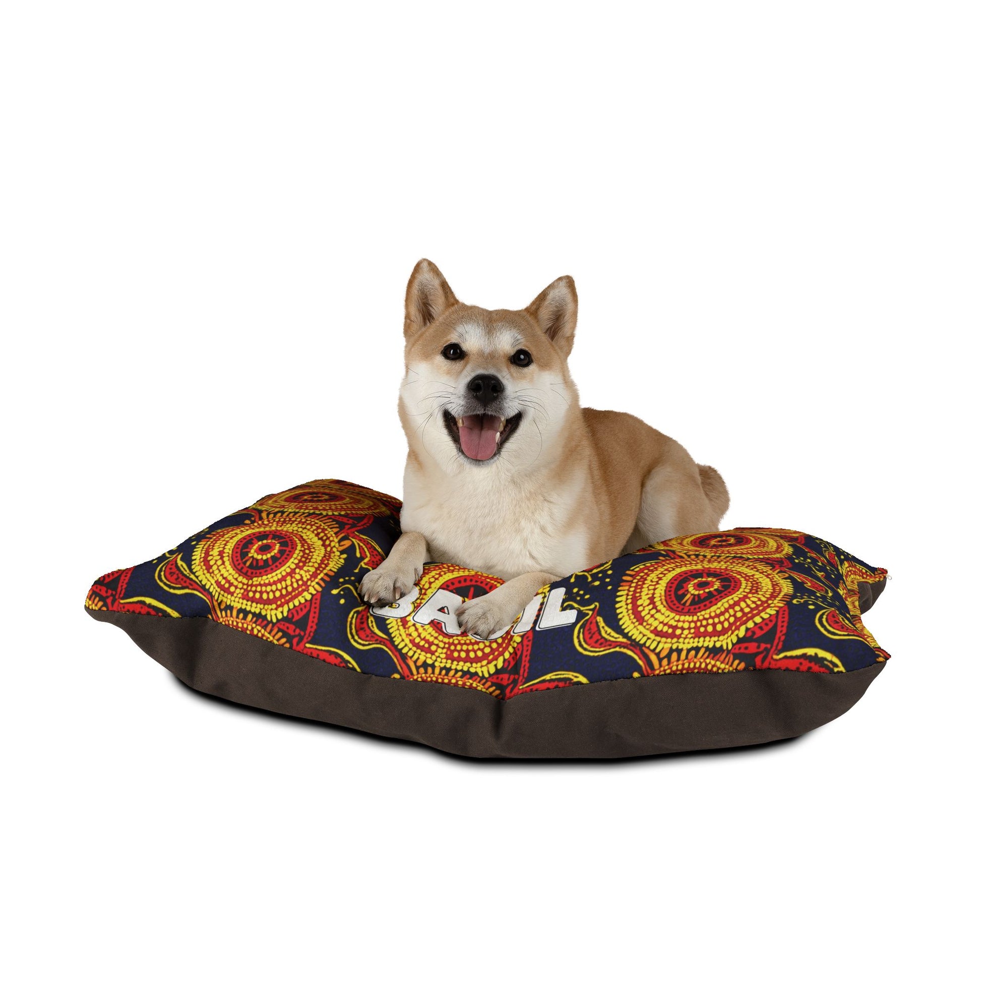 A Shiba Inu dog with tan and white fur lies on an African-inspired cushion featuring intricate red, yellow, and black patterns. The dog looks happy with its mouth open and tongue out, against a plain white background.