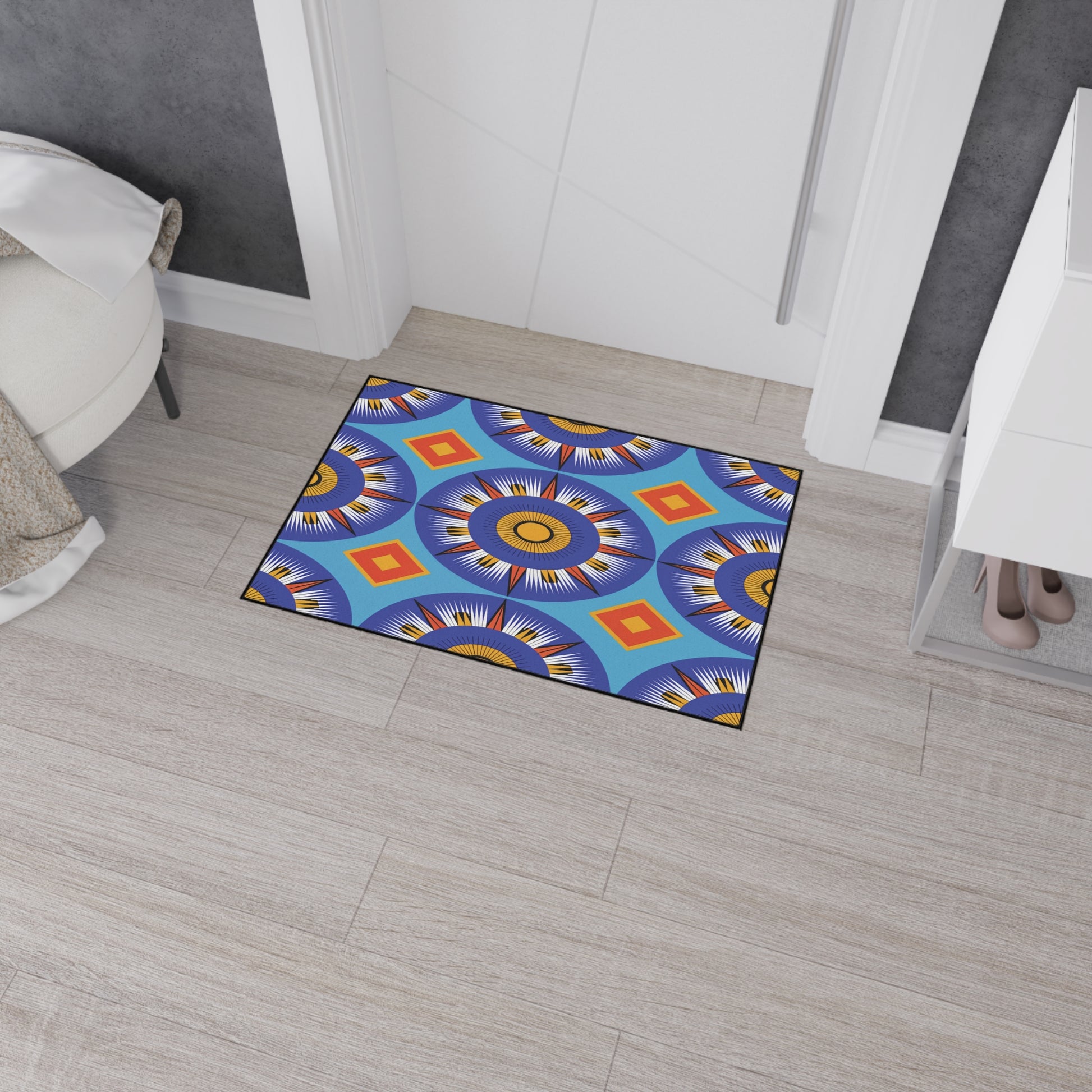 A colorful doormat with bold blue, orange, yellow, and red geometric sunburst patterns inspired by African-inspired decor lies on a light wood floor. It features non-slip rubber backing and sits near white walls, a shoe cabinet, and a bed with a white blanket.