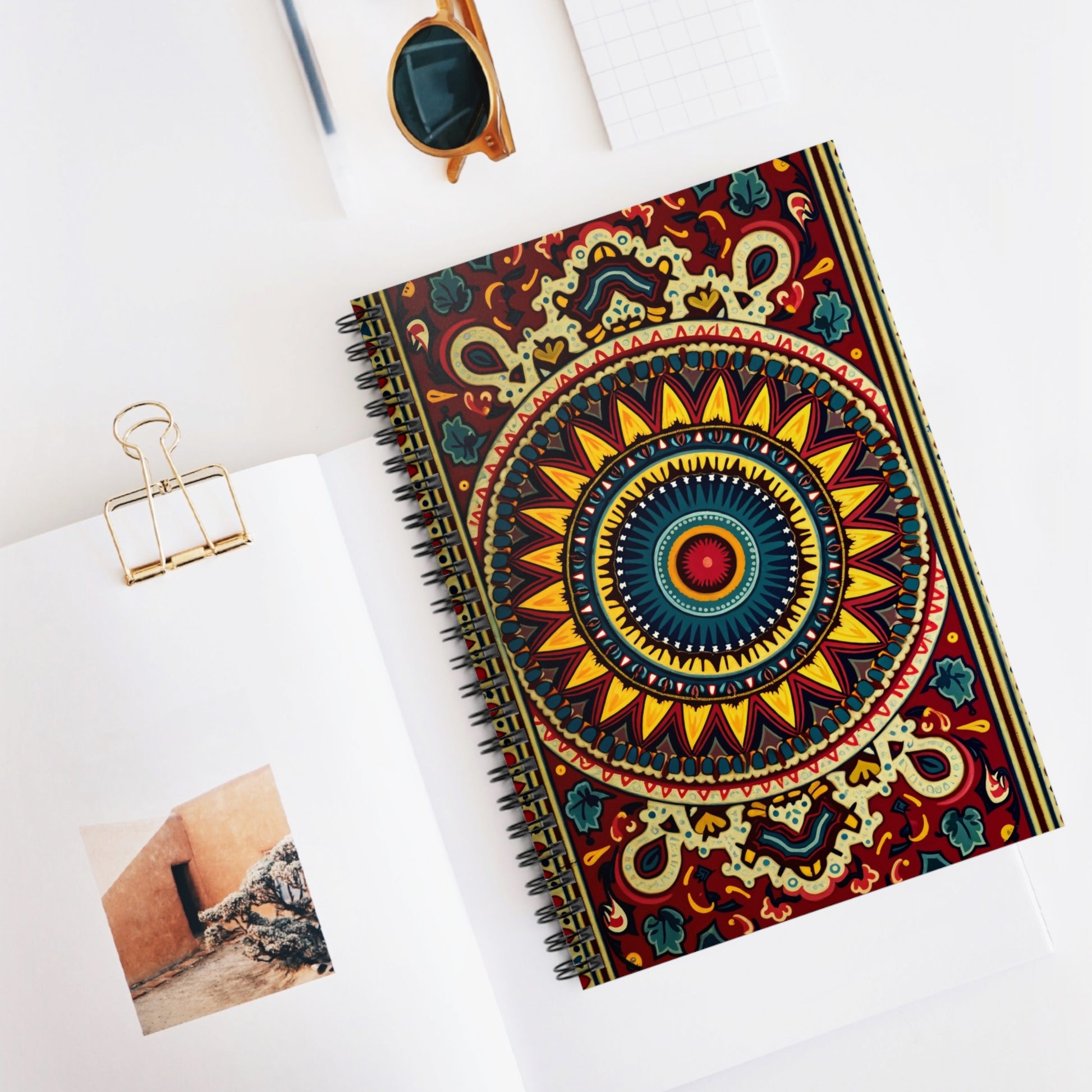 A culturally inspired notebook with a vibrant, colorful mandala design graces a white desk. Nearby lie an open book with a photo, sunglasses, a metal paper clip, and a small gridded notepad, all neatly arranged.