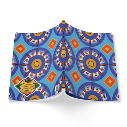 A book with a blue cover features a vibrant, symmetrical pattern of circular sunburst shapes in blue, yellow, and white, accented by orange diamonds. A gold sticker on the bottom left reads “AFRO COMFORT SHOP.” This African-inspired journal is open and viewed from above.
