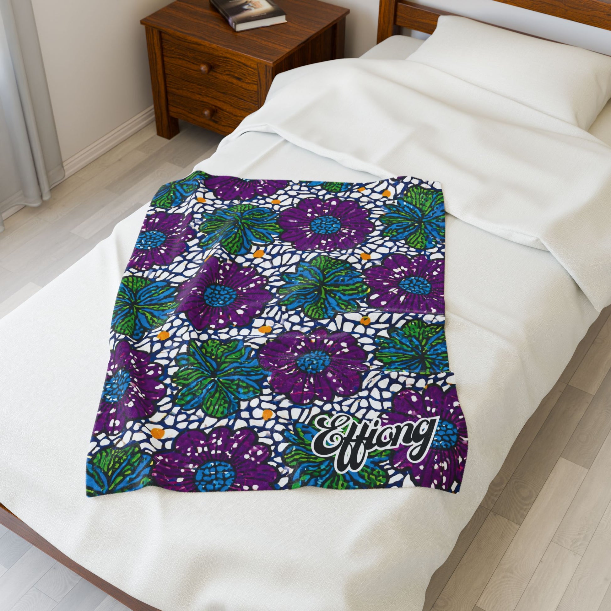 A colorful African-inspired blanket with a vibrant floral pattern in purple, green, blue, and white lies on a neatly made bed. Near the corner, “Effiong” is written in cursive, making it the perfect personalized velveteen blanket for any space.
