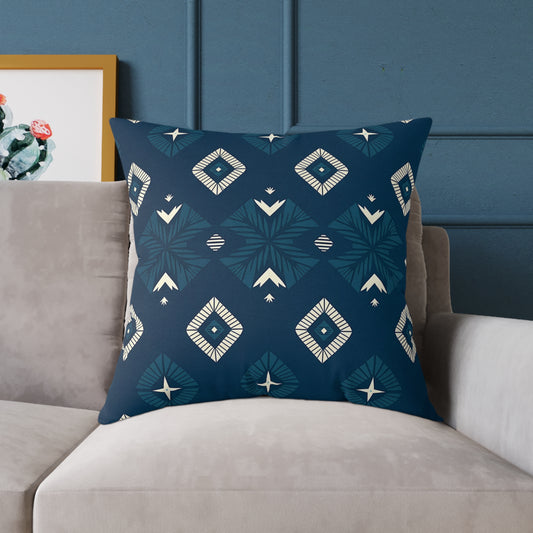 A vibrant throw pillow in navy blue with geometric white and teal diamond patterns rests on a light gray sofa. The background wall is blue with vertical molding, and a framed cactus artwork is partially visible behind the pillow on the left.