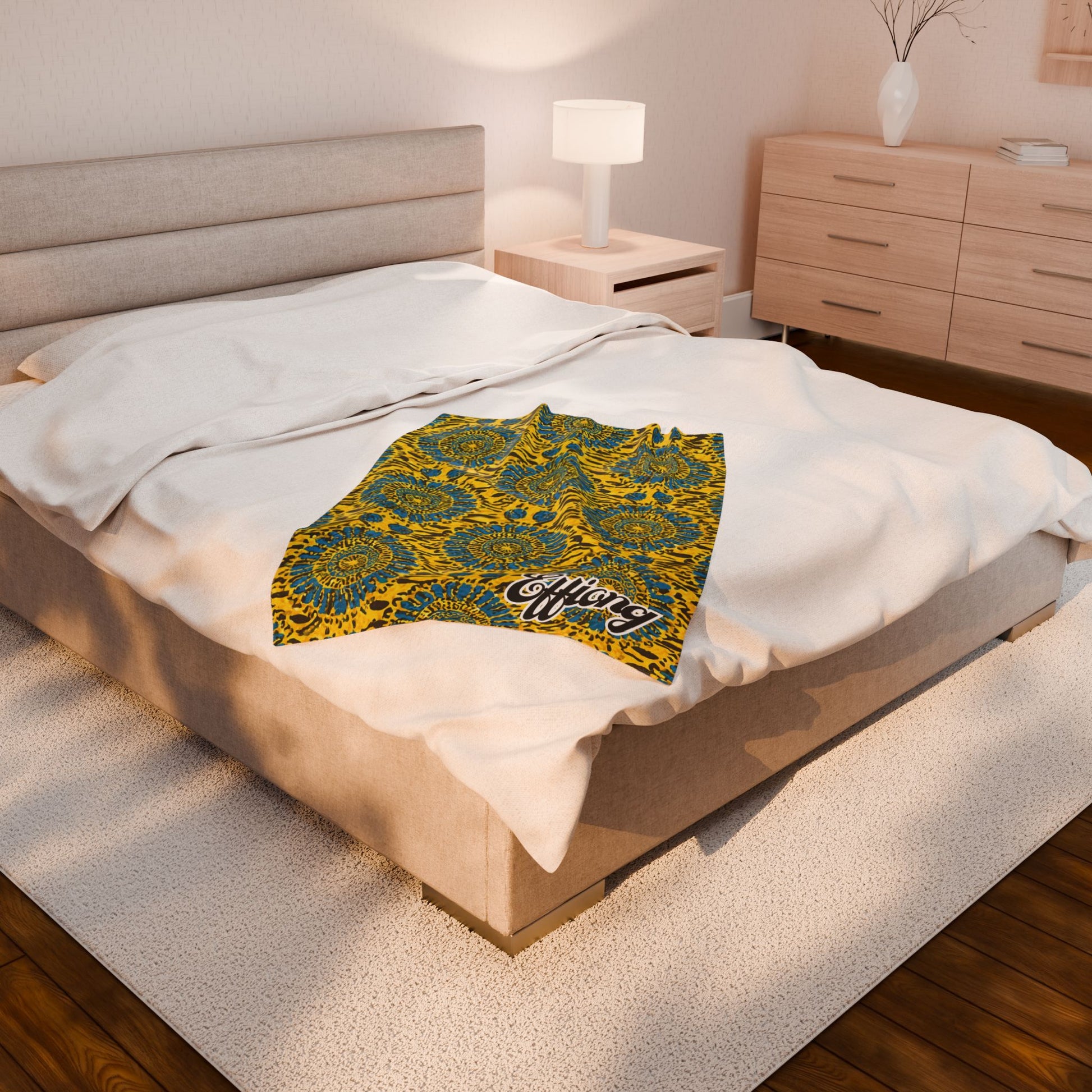 A modern bedroom with a beige upholstered bed, white bedding, and an African-inspired blanket in vibrant yellow, blue, and green draped on top. A nightstand with a lamp and a wooden dresser with a white vase complete the space.