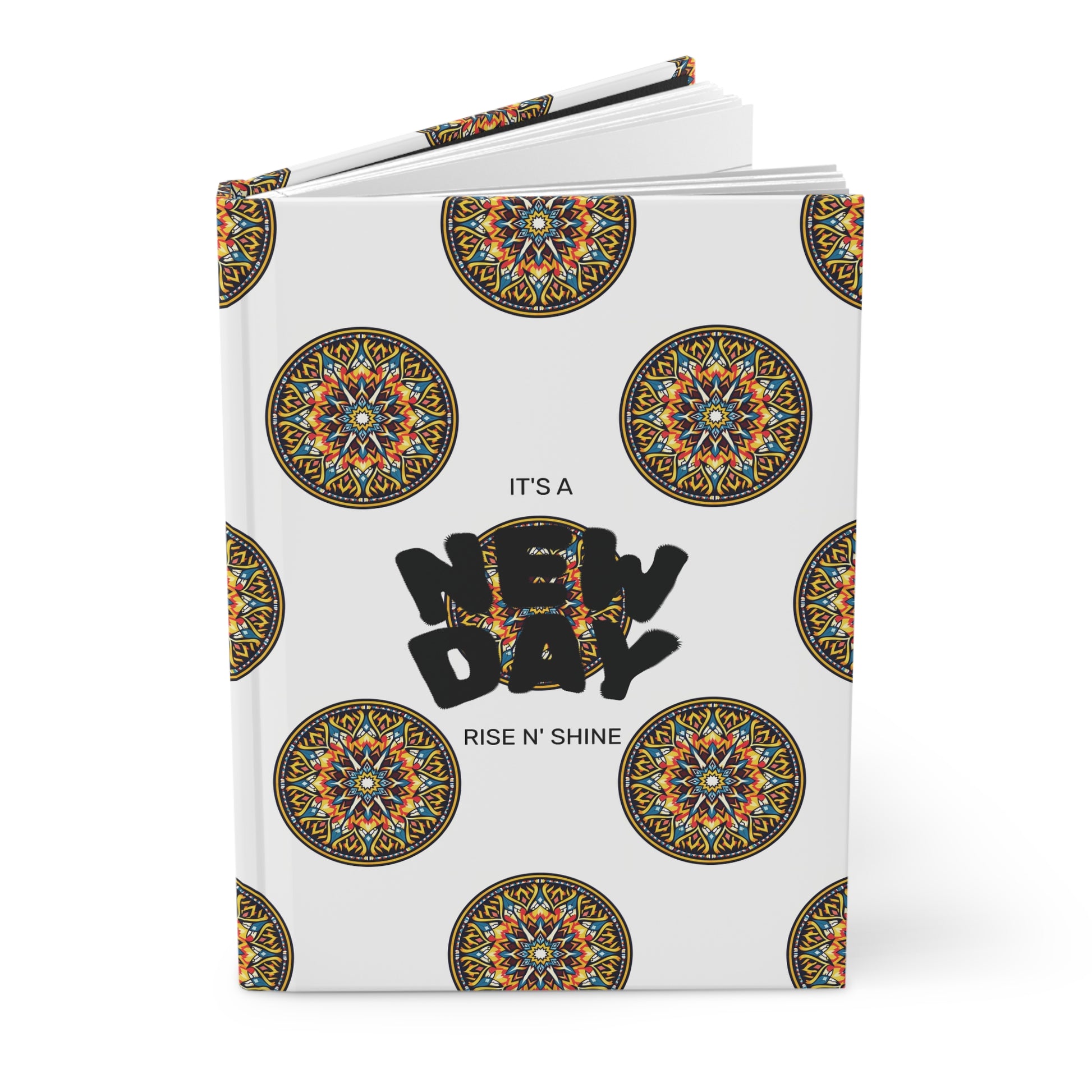 A hardcover journal with a white cover decorated with colorful circular mandala patterns. Centered bold black text reads “NEW DAY,” with smaller text above and below stating “IT’S A” and “RISE N’ SHINE.” Perfect for your wellness ritual, the notebook is slightly open.
