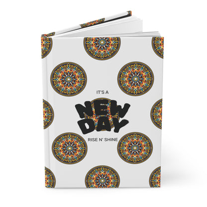 A hardcover journal with a white cover decorated with colorful circular mandala patterns. Centered bold black text reads “NEW DAY,” with smaller text above and below stating “IT’S A” and “RISE N’ SHINE.” Perfect for your wellness ritual, the notebook is slightly open.