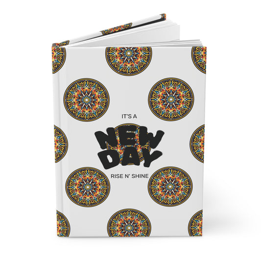 A hardcover journal with a white cover decorated with colorful circular mandala patterns. Centered bold black text reads “NEW DAY,” with smaller text above and below stating “IT’S A” and “RISE N’ SHINE.” Perfect for your wellness ritual, the notebook is slightly open.