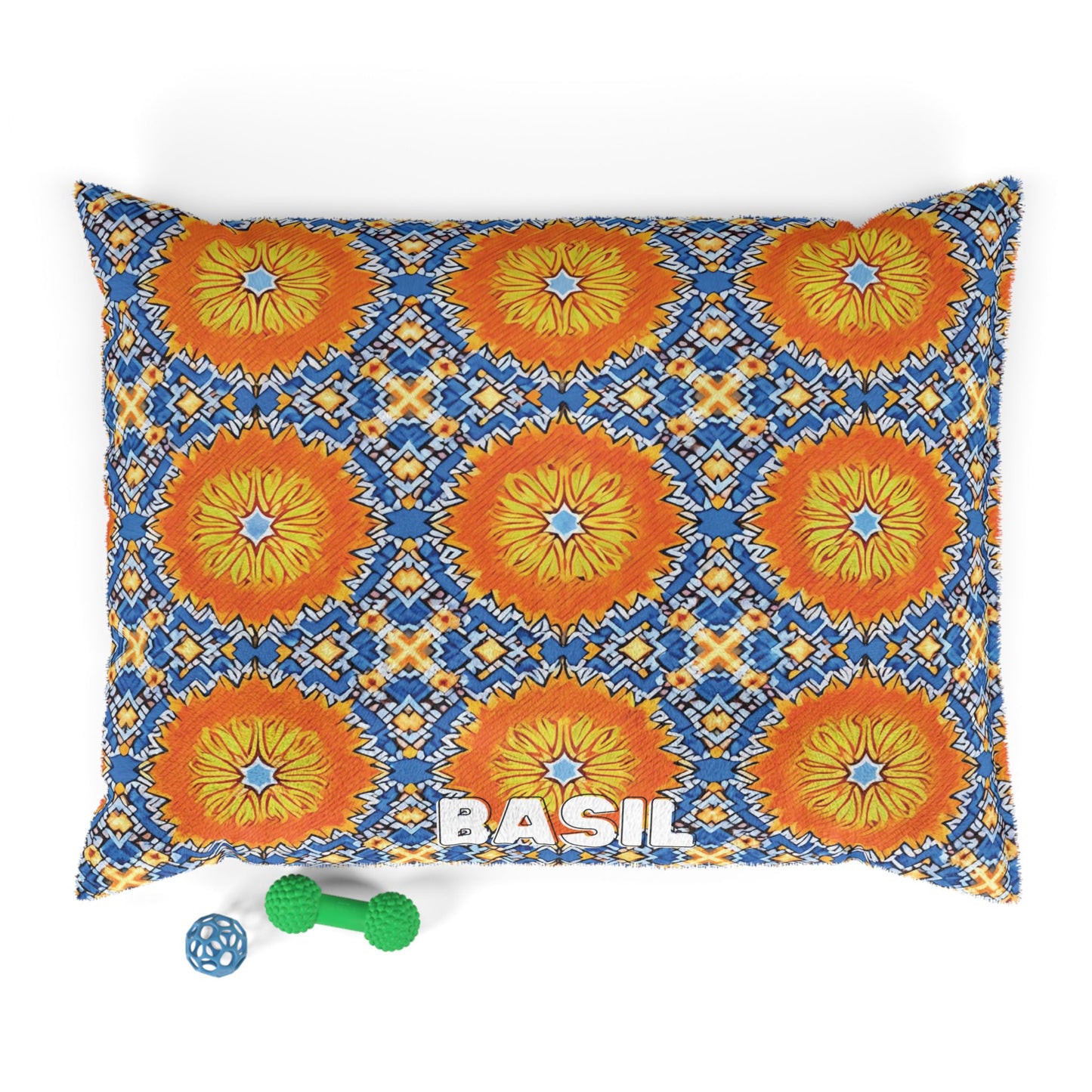 A stylish pet bed with a colorful pattern of orange, yellow, and blue geometric floral designs features the name BASIL in white letters. Next to this cozy pet pillow are two toys: a small blue ball and a green dumbbell-shaped chew toy.