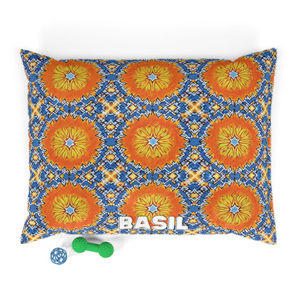 A stylish pet bed with a colorful pattern of orange, yellow, and blue geometric floral designs features the name BASIL in white letters. Next to this cozy pet pillow are two toys: a small blue ball and a green dumbbell-shaped chew toy.